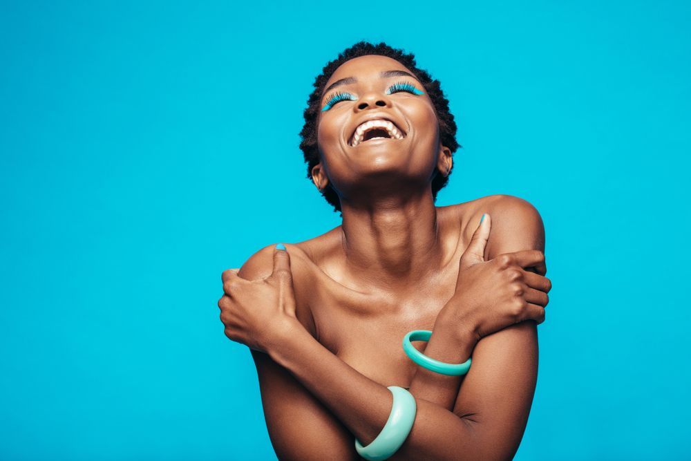 Cheerful african woman hugging herself against blue background. Female model wearing vibrant makeup laughing in studio.