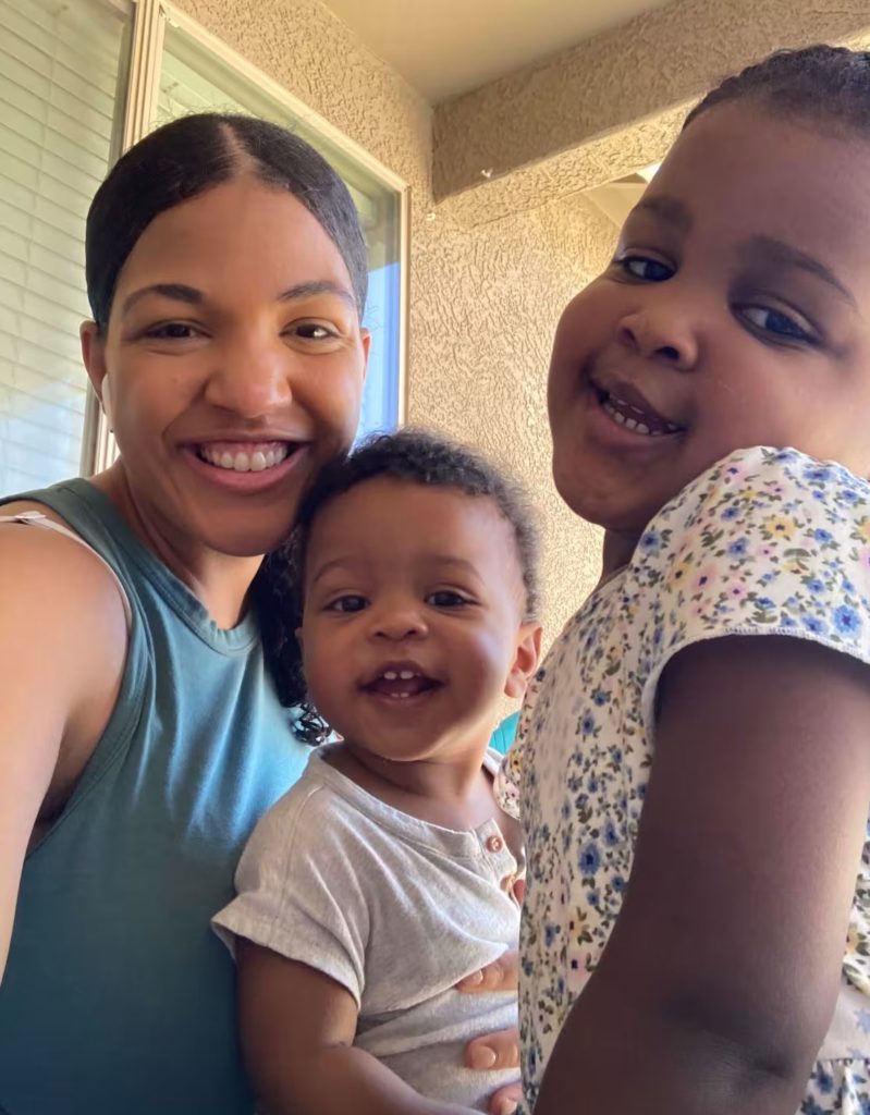 A mom smiles with her 2 kids. Window and wall in the background.