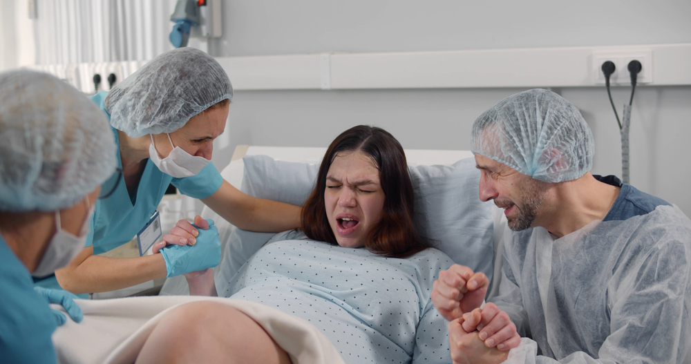 Husband and nurses supporting pregnant woman hardly breathing during child birth. Young lady delivering baby in hospital ward with husband and doctors help