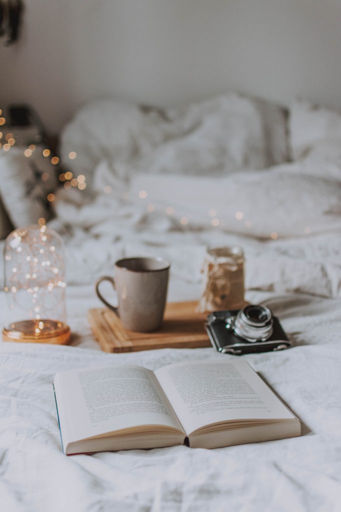 A cozy looking bed with fairy lights, an open book and beverage and a camera. Blurred comforter in the background.