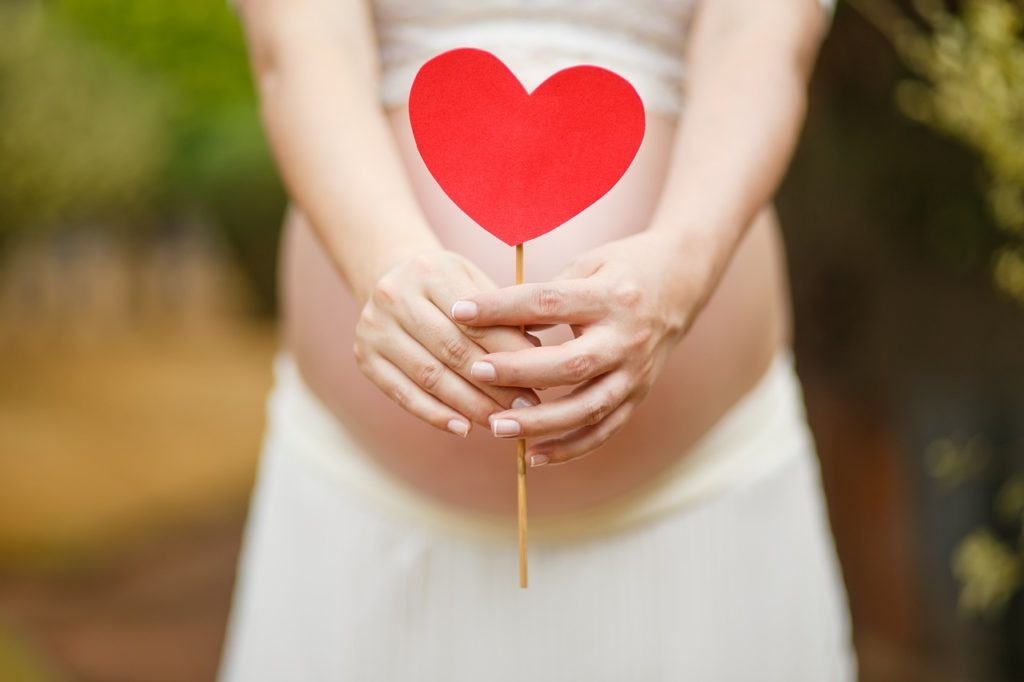 A pregnant woman holds a heart in front of her.