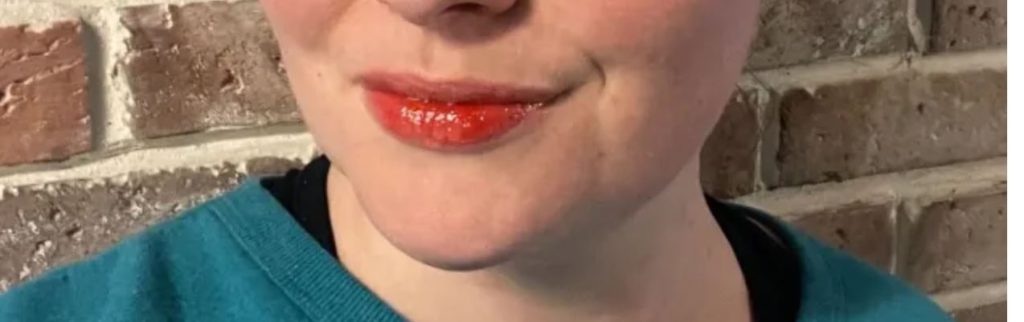 A woman with bright red lips wearing a teal/dark green or blue shirt. A brick background.