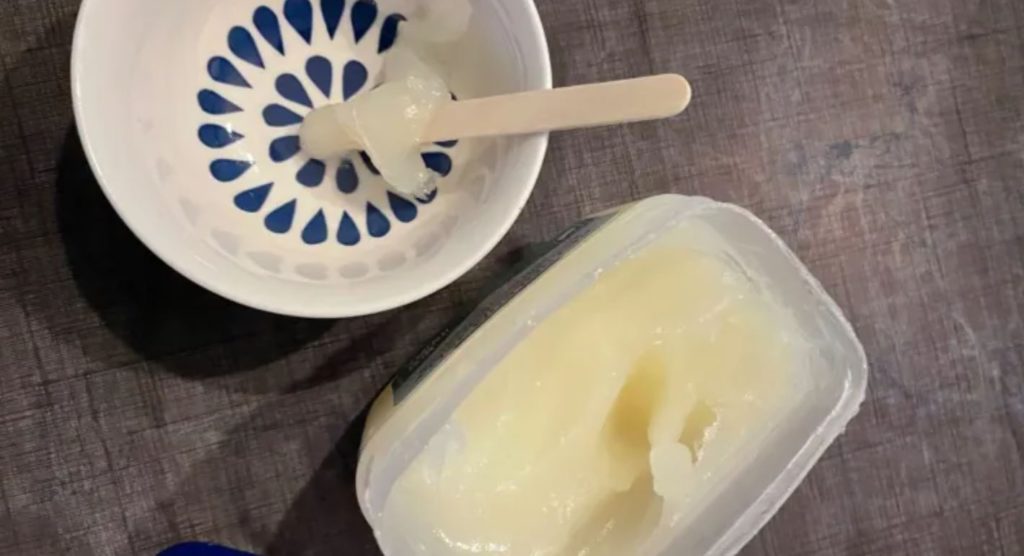 A tub of Vaseline next to a bowl with a popsicle stick for scooping. A wooden background.