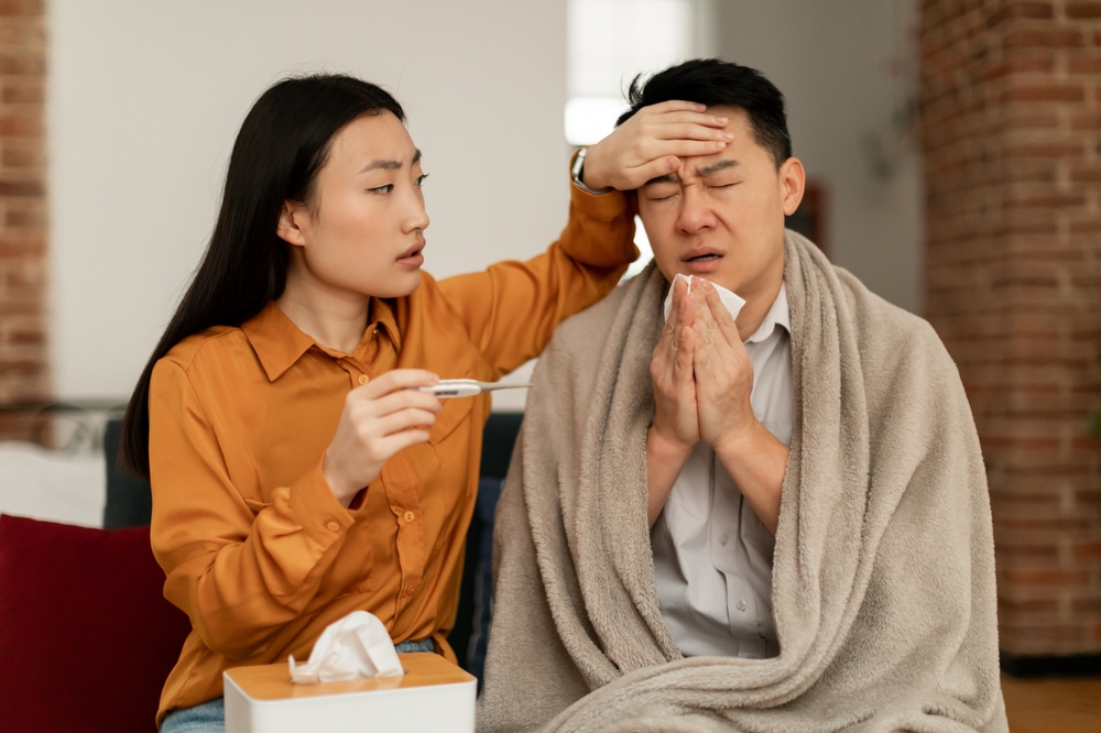 Asian woman treating sick husband, holding thermometer and touching man forehead, male having seasonal flu and cold, sitting on couch at home. Fever, influenza treatment concept