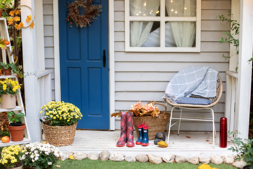 Cozy Autumn terrace with chair, plaid, rubber boots, baskets with chrysanthemums and pumpkins. Decorations in autumn patio for relax. Stylish fall decor on front porch home. Autumn wooden porch home
