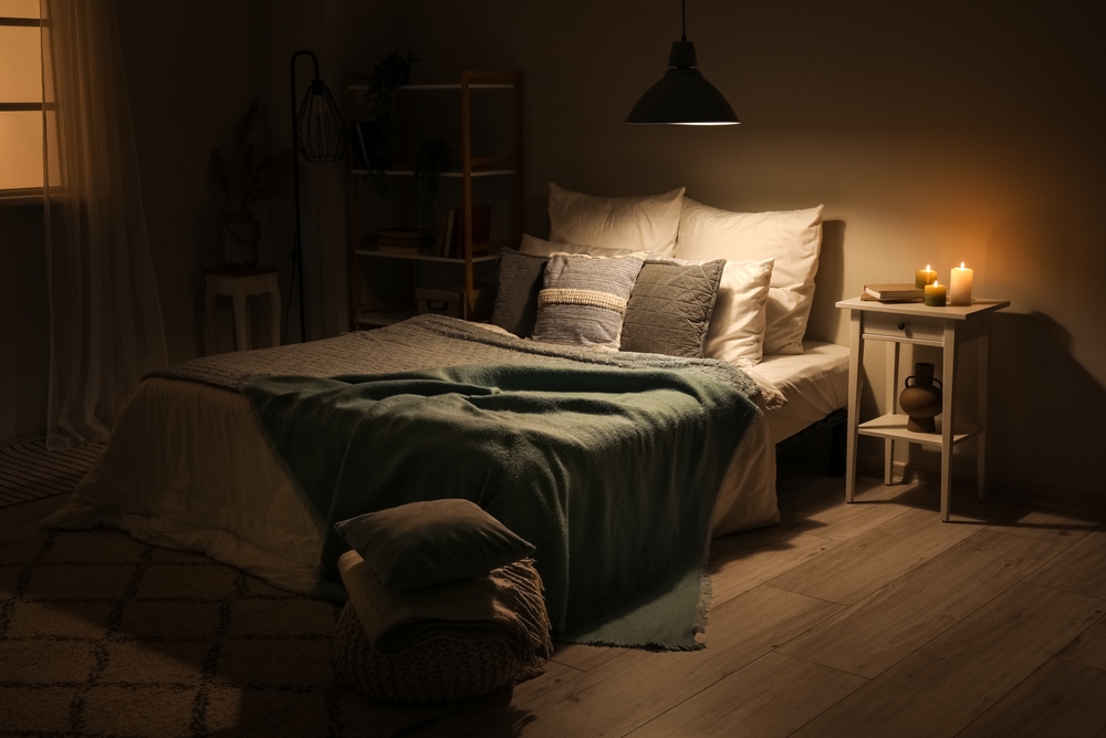Interior of bedroom with green blankets on bed, burning candles and glowing lamp at night