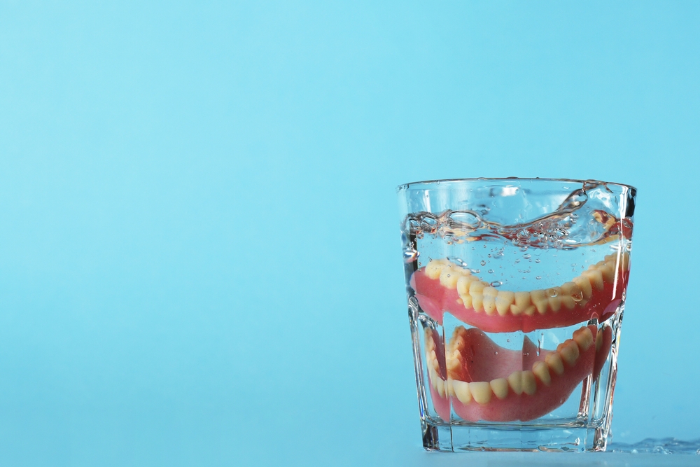 Dentures in a glass of water, blue background with copy space. Dental prosthesis health care. False teeth, dentures.

