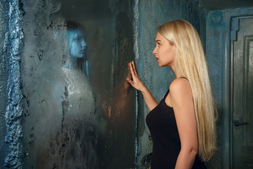 Frightened young woman looking in the mirror and seeing in reflection a ghost girl
