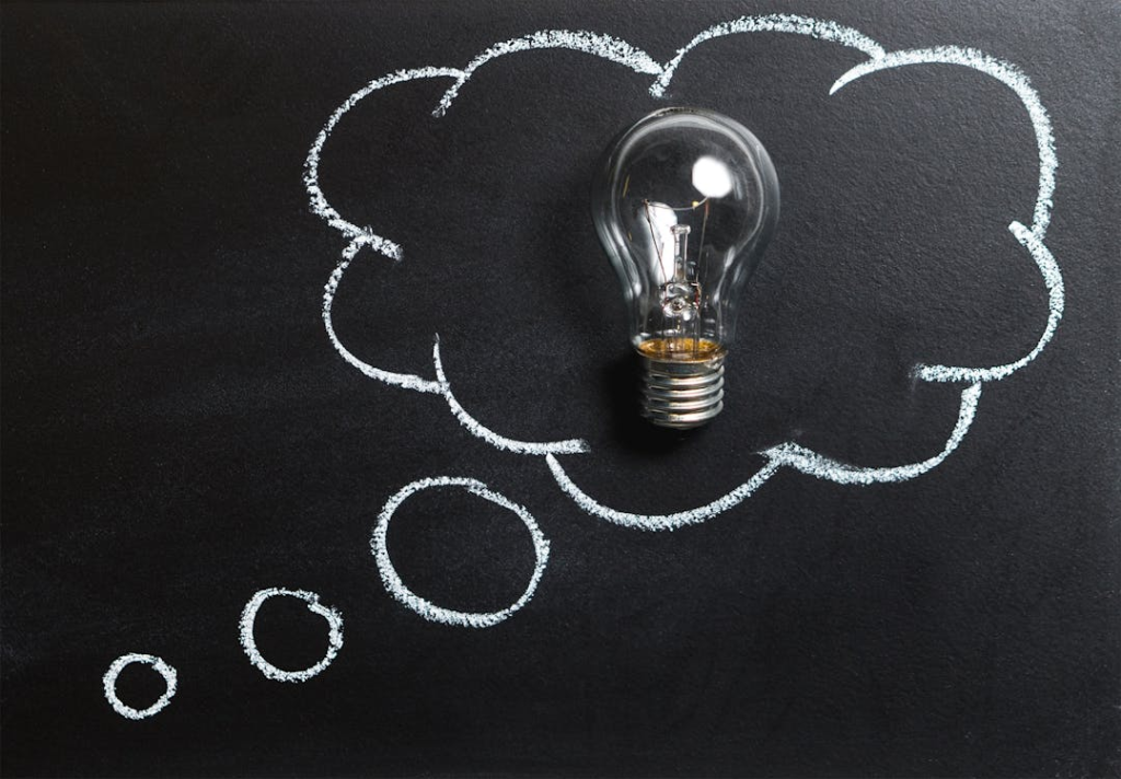 A clear incandescent light bulb placed inside a thought bubble drawn with white chalk on a black chalkboard. The thought bubble is connected to smaller circles below it, resembling the classic cartoon representation of thinking. The contrast between the three-dimensional light bulb and the hand-drawn chalk illustration symbolizes ideas, inspiration, or creative thinking.