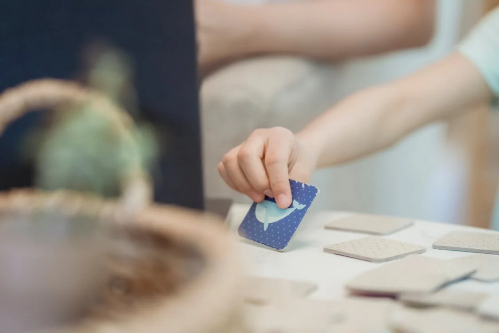 A close-up photograph of a hand picking up a small blue card with a white whale illustration on it from a table. Several other plain white square cards are visible on the surface. The image is taken from a shallow angle with a soft focus on the background, where the outline of what appears to be a plant or basket can be seen.