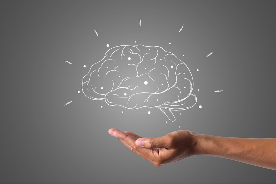 A digital illustration of a white outlined brain floating above an outstretched hand against a gray background. The brain drawing features intricate details of neural folds and is surrounded by small white dots and short lines suggesting activity or energy. The hand appears to be presenting or supporting the glowing brain concept.