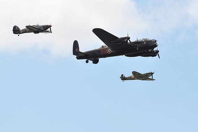 Three historic military aircraft flying in formation against a partly cloudy blue sky. A large four-engine Lancaster bomber is prominently featured in the center, flanked by a Hurricane fighter plane on the left and a Spitfire fighter on the right. The aircraft display traditional RAF roundel markings, suggesting this is likely a commemorative flight or airshow of World War II era planes.