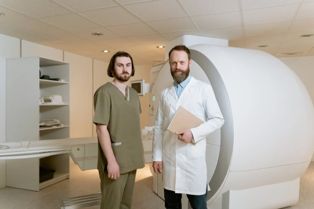 Two healthcare professionals standing in a medical imaging room next to an MRI machine. One person wears a sage green scrub uniform while the other wears a white lab coat over blue clothing and holds a folder. They are positioned beside the large white cylindrical MRI scanner with its characteristic tunnel opening. The clinical space features white cabinets and drop ceiling tiles with recessed lighting.