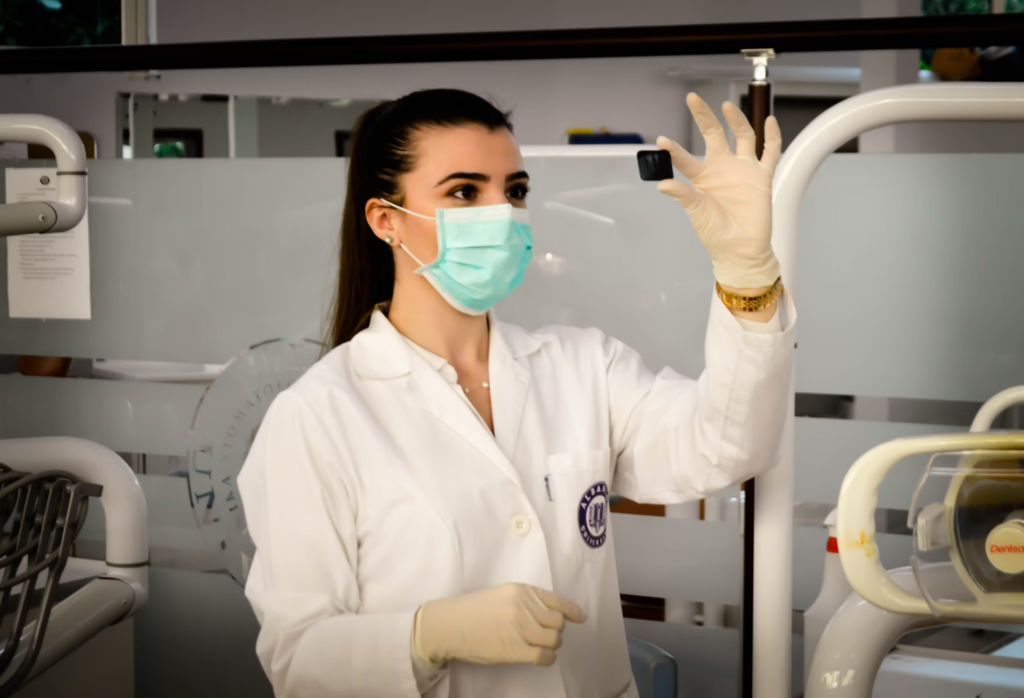 Female healthcare professional in a white lab coat and surgical mask examining a medical vial or specimen. She is wearing latex gloves and standing in a clinical laboratory or medical setting with equipment visible in the background.