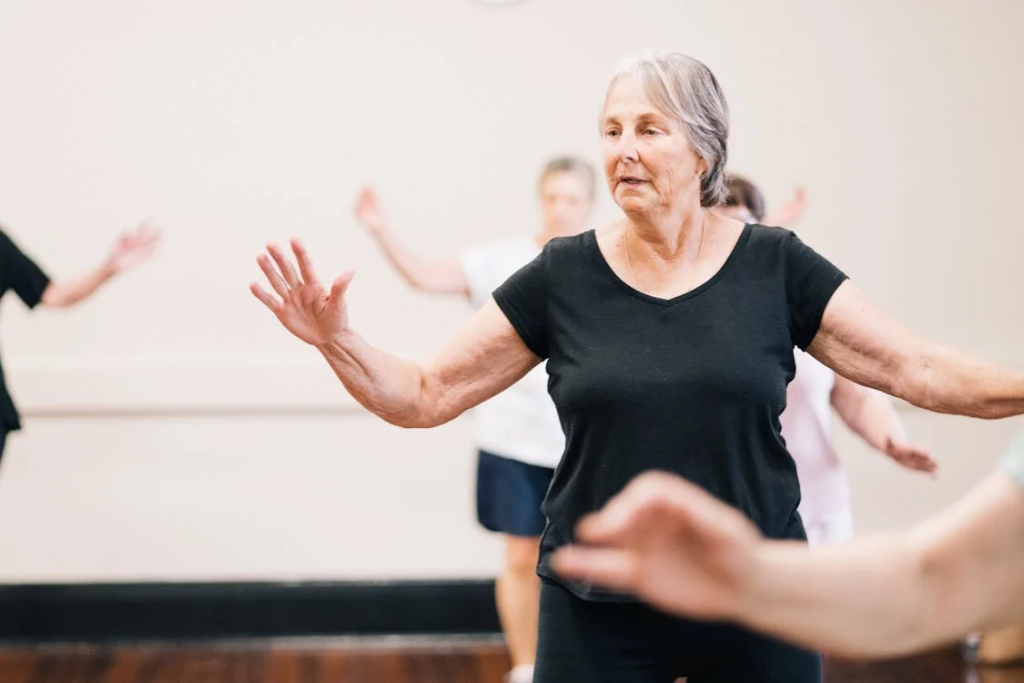 An older woman with silver hair participating in a group exercise class. She is wearing a black t-shirt and has her arms outstretched while practicing balance exercises or gentle movement in a studio setting.