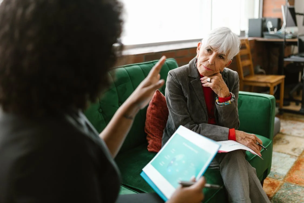 An older woman in a grey blazer listening to a healthcare advisor during a consultation.