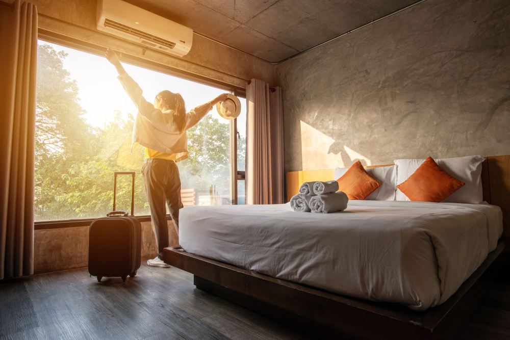 Portrait of tourist woman raised her hands and standing nearly window, looking to beautiful view with her luggage in hotel bedroom after check-in. Conceptual of travel and vacation.