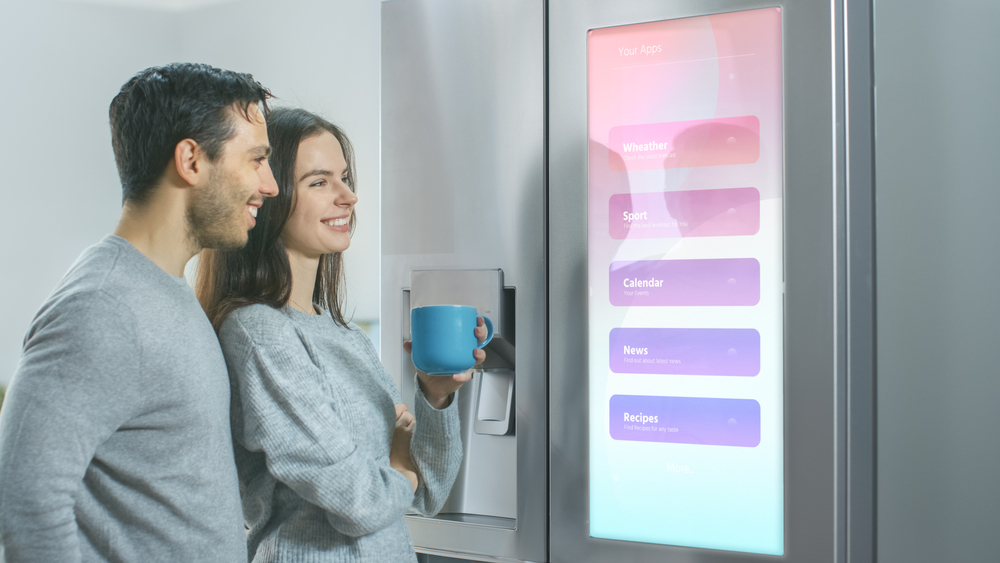 Beautiful Young Couple Drink Morning Coffee in the Kitchen. They Check the Weather Forecast and a To Do List on a Smart Fridge at Home. Apartment is Bright and Cozy.

