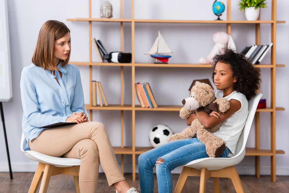 Psychologist talking to frustrated african american girl hugging teddy bear during consultation in office