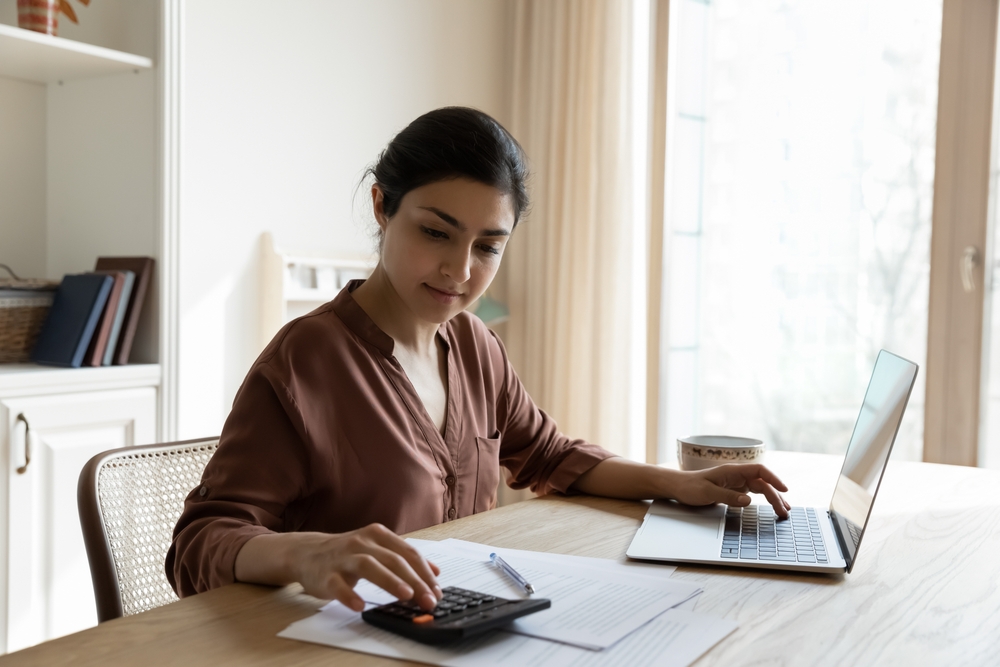 Household budget. Young Indian female sit at table calculate utility payments before make money transfer in online bank app on laptop. Focused millennial business woman pay taxes plan personal