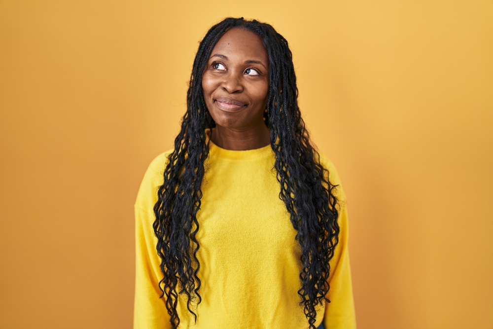 African woman standing over yellow background smiling looking to the side and staring away thinking.