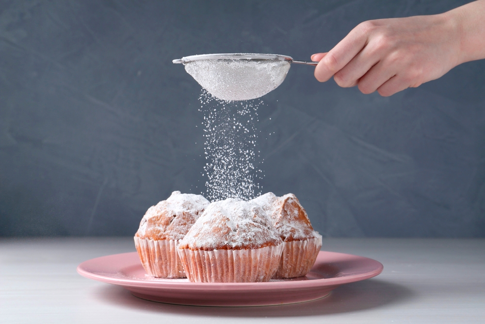 Woman with sieve sprinkling powdered sugar onto muffins at white wooden table, closeup
