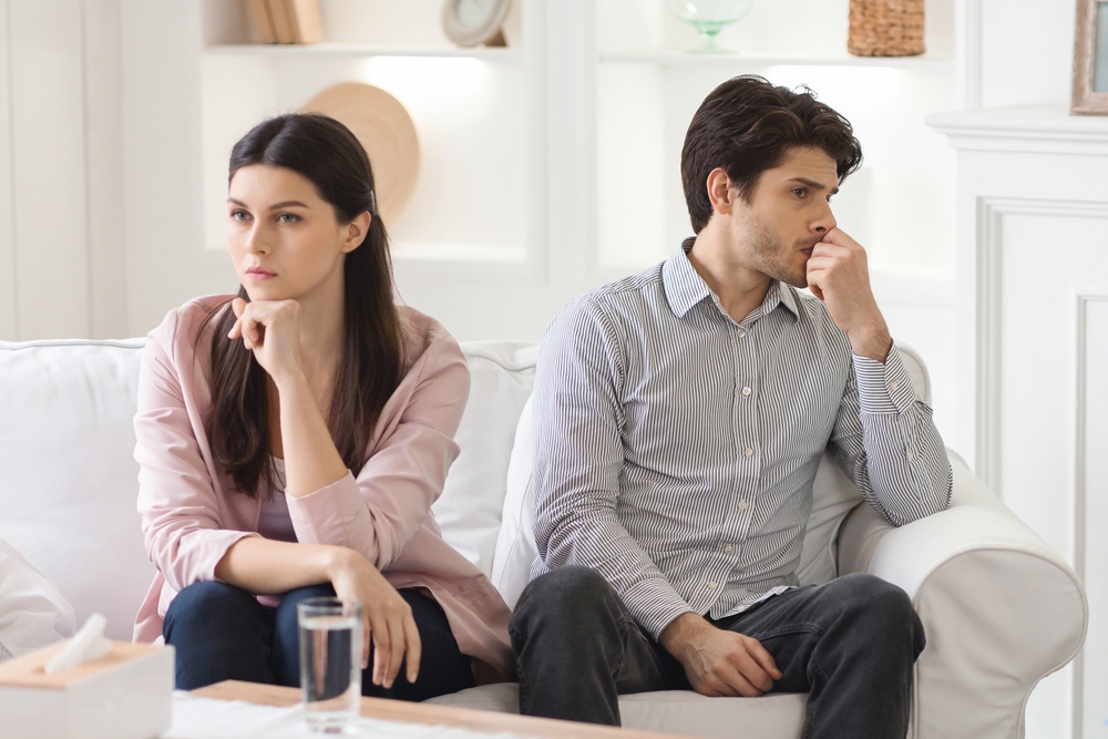 A young couple sits on a sofa in a brightly lit living room. Both appear to be lost in thought, staring off into the distance with serious expressions. Crisis in relationships
