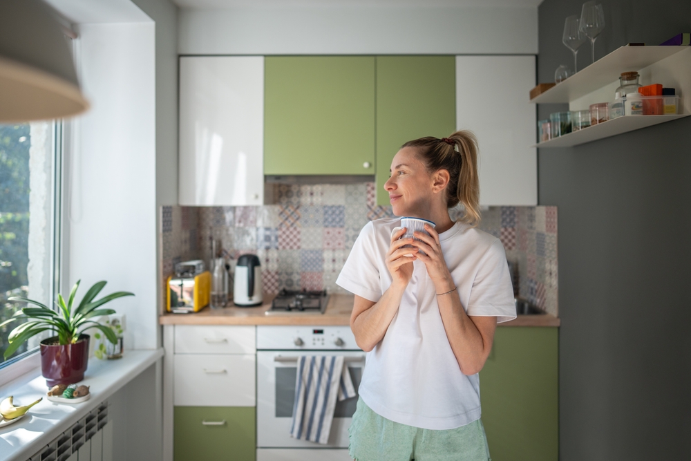 Joyful smiling woman with morning coffee, observing outside from domestic kitchen. Slow morning, contentment, relaxed mood, no rush weekend, mindful slow living, refreshing break, cozy routine.