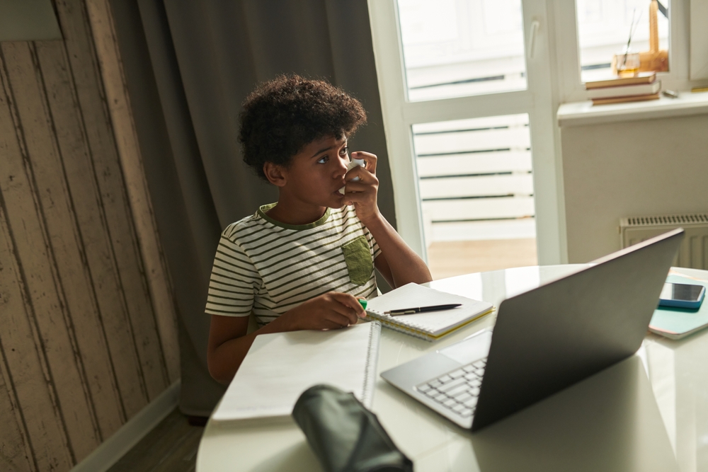 Modern diligent schoolboy with asthma pumping inhaler during attack while sitting by table in front of laptop during online lesson