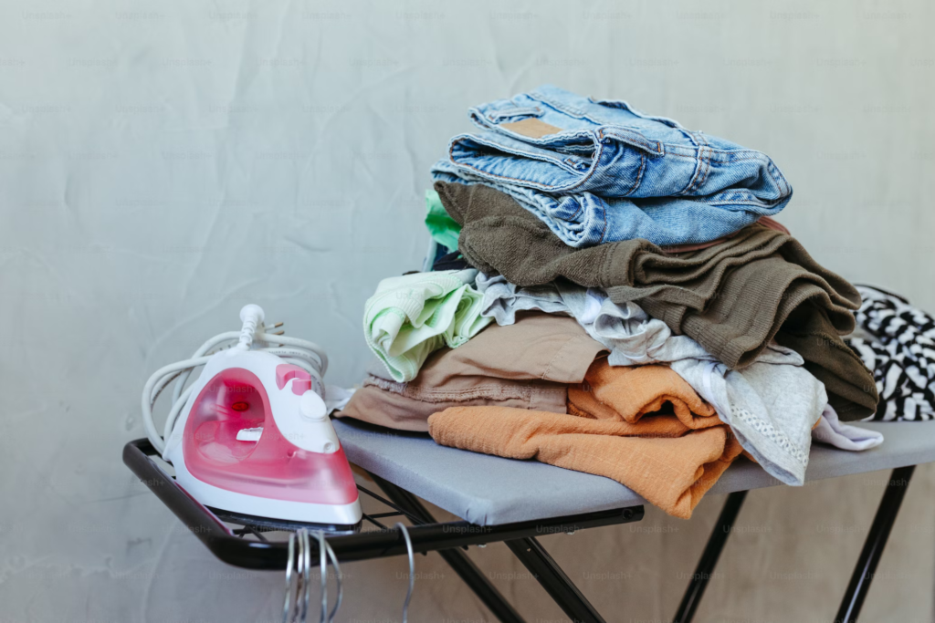 Pink iron on an ironing board next to a stack of wrinkled clothes waiting to be pressed, illustrating high-maintenance garments.