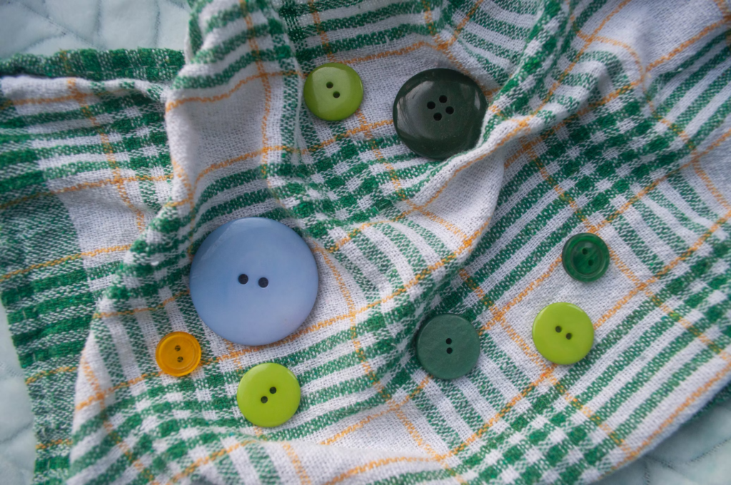 Close-up of green, blue, and yellow buttons scattered on a plaid fabric, representing clothing items awaiting repair.