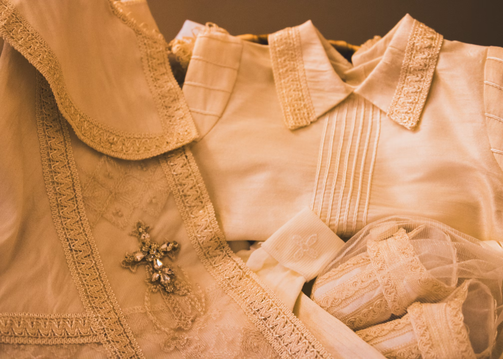 Vintage cream-colored clothing items with lace details and embellishments stacked together, representing sentimental pieces that are difficult to part with.