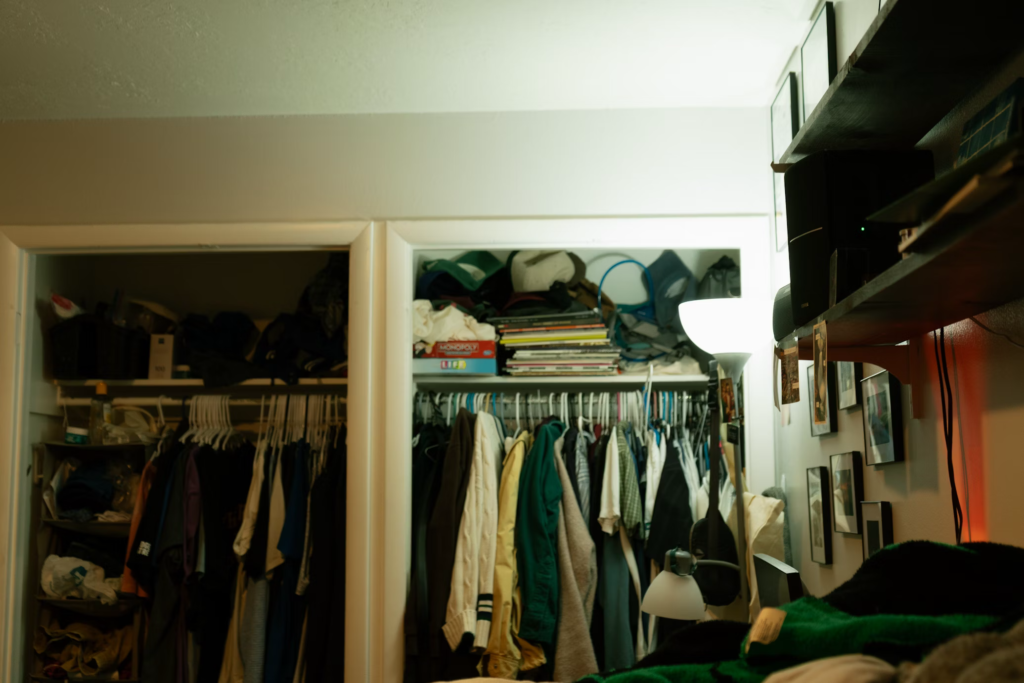 Cluttered closet with clothing, books, and miscellaneous items packed tightly on shelves and hanging rods, showing seasonal overflow.