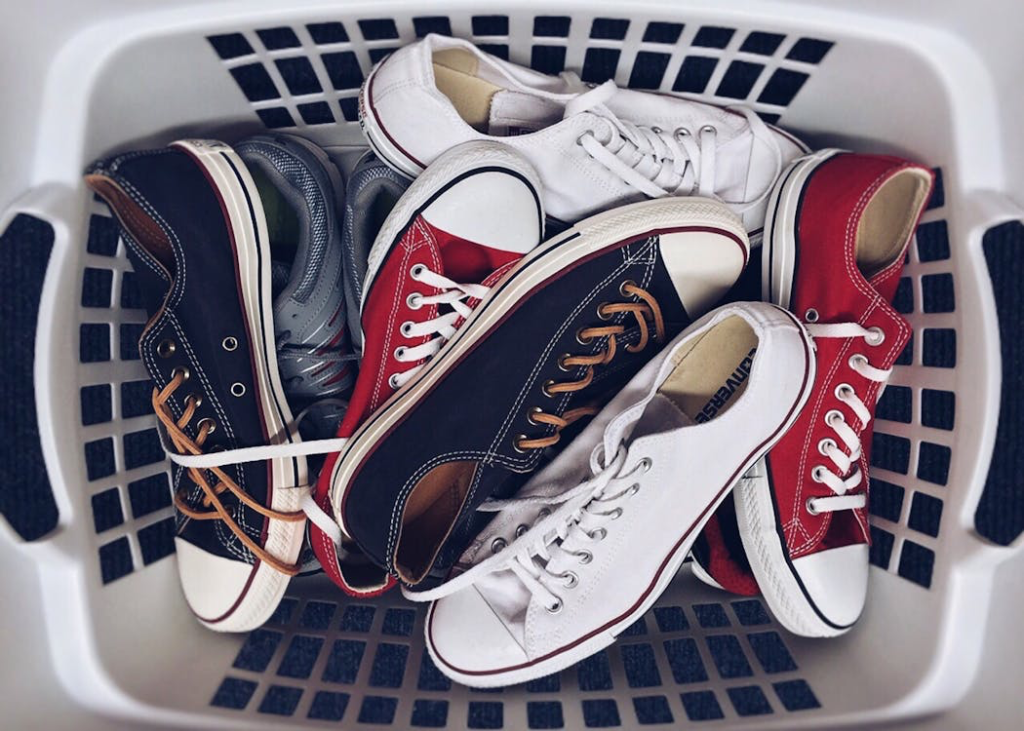 White laundry basket filled with multiple pairs of sneakers in red, white, blue, and black, demonstrating an excess of casual footwear.