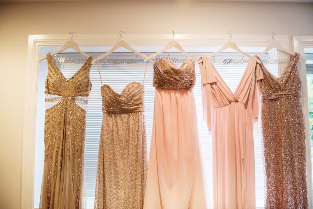 Five formal gowns in gold, champagne, and blush tones hanging on personalized hangers against a window, representing rarely worn formal attire.