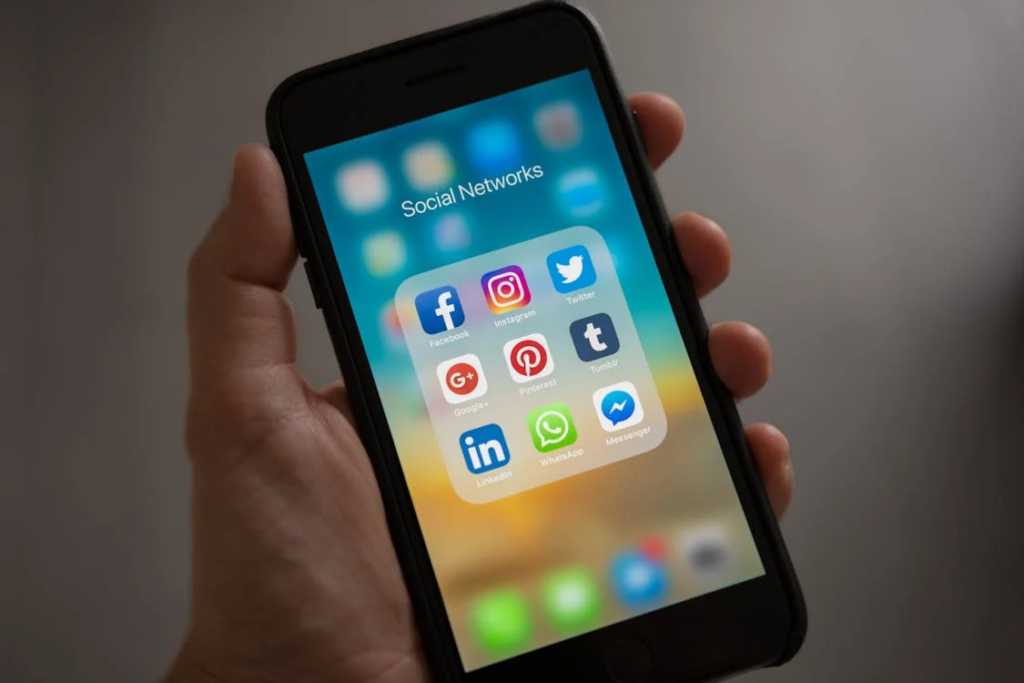 Close-up of a hand holding a smartphone displaying a folder of social media application icons including Instagram, Facebook, Twitter, Pinterest, LinkedIn, WhatsApp, and Messenger against a blurred background.