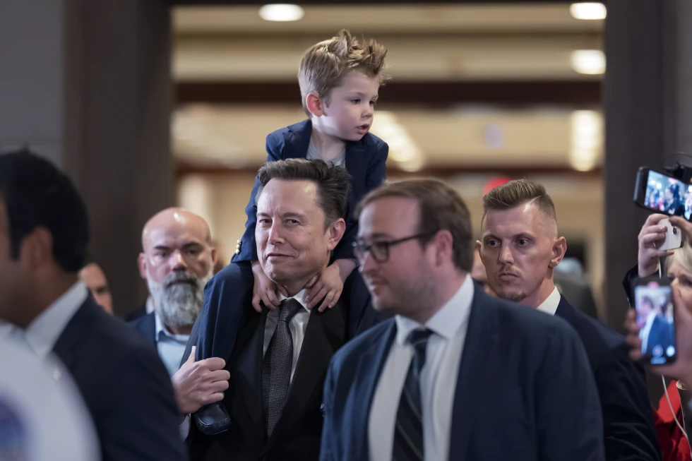 Elon Musk with his youngest son on his shoulders, surrounded by security and reporters.