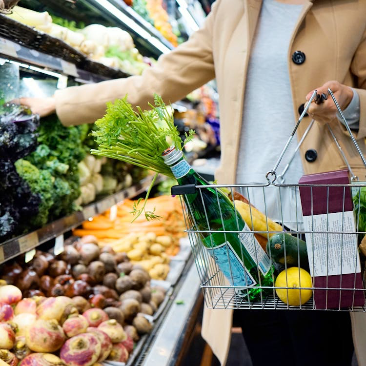 Shopper in a beige coat selecting fresh herbs while shopping, holding a metal basket filled with produce and other items.