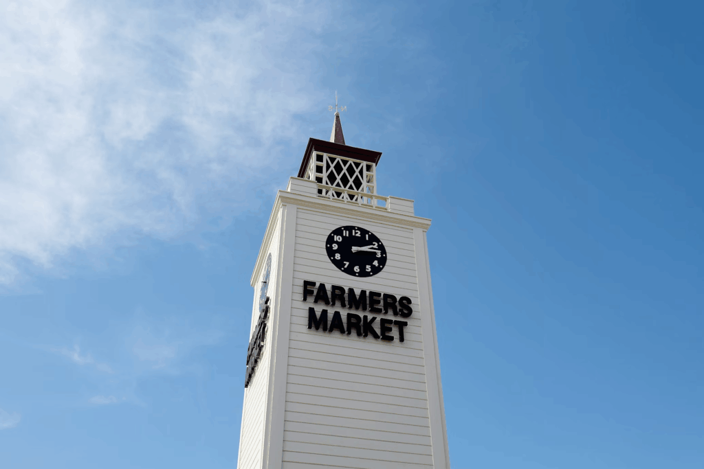 White clock tower with 'FARMERS MARKET' sign against a blue sky, marking a popular shopping destination as consumers search for alternatives to high grocery prices.