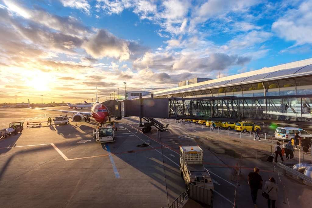 An airport terminal at sunset or sunrise with beautiful cloudy sky. The image shows an airplane connected to a jet bridge, with ground crew vehicles and airport operations visible on the tarmac. The golden light creates a dramatic atmosphere of travel.