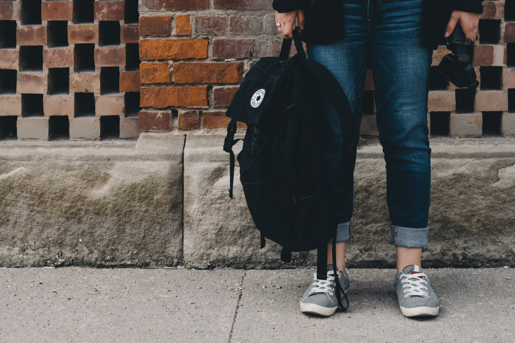 A person in blue jeans and gray sneakers holding a black backpack. The photo is cropped to show only the lower half of the person standing against a brick wall, highlighting the backpack as a personal item for travel.