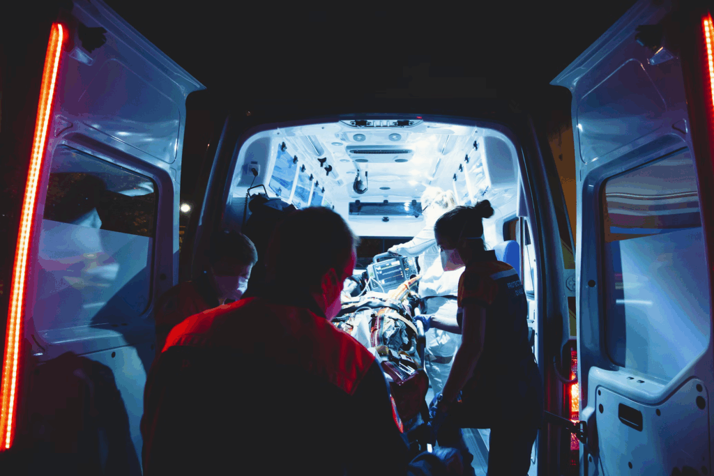 Emergency medical technicians loading a patient into an ambulance at night. The image shows the back doors of an ambulance open with its interior illuminated by blue lights. Medical personnel in silhouette are visible attending to what appears to be a stretcher. The dramatic lighting emphasizes the urgency of the emergency response situation.