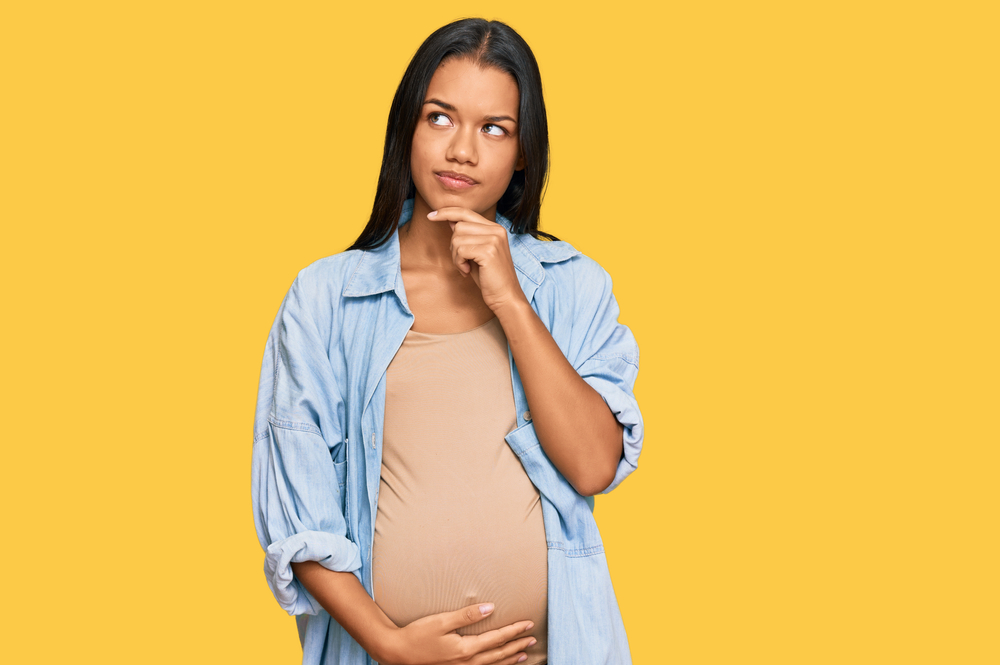 Beautiful hispanic woman expecting a baby showing pregnant belly serious face thinking about question with hand on chin, thoughtful about confusing idea
