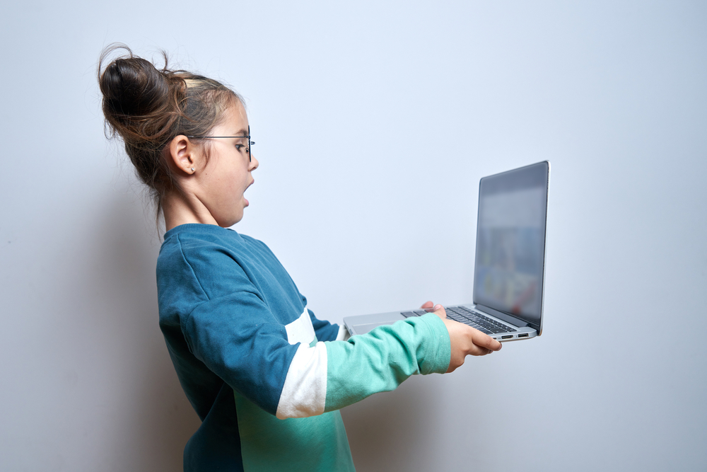 Shocked child girl with open mouth and bulging eyes looks at a computer laptop screen. Internet porn censorship concept, adult content 18+