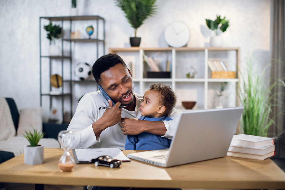 Afro american man talking on mobile, working on laptop and carrying baby son. Male freelancer sitting at home office and using modern gadgets.
