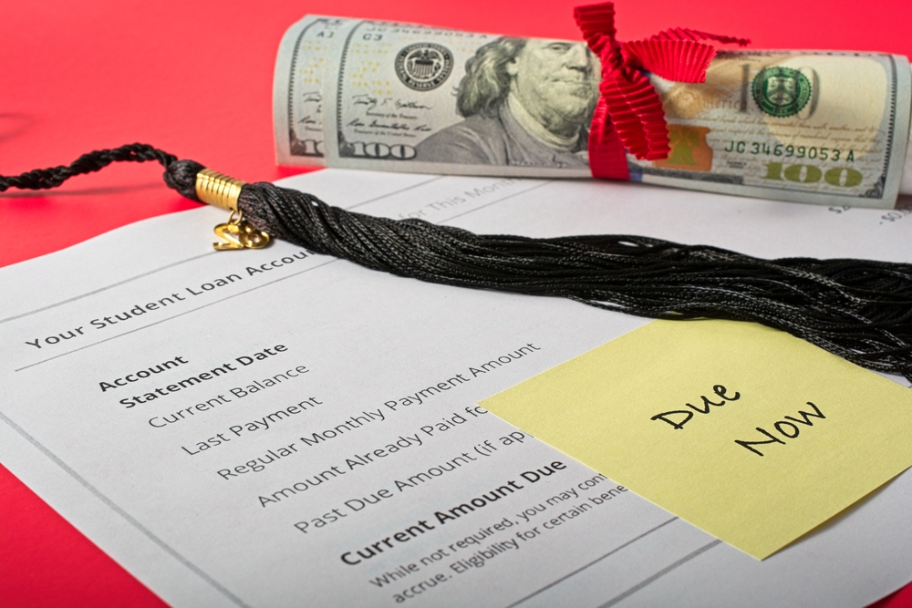 Post-it labeled ‘Due Now’ affixed to student loan invoice under black graduation cap tassel and 100 dollar bill wrapped diploma scroll. Red ribbon and red background.