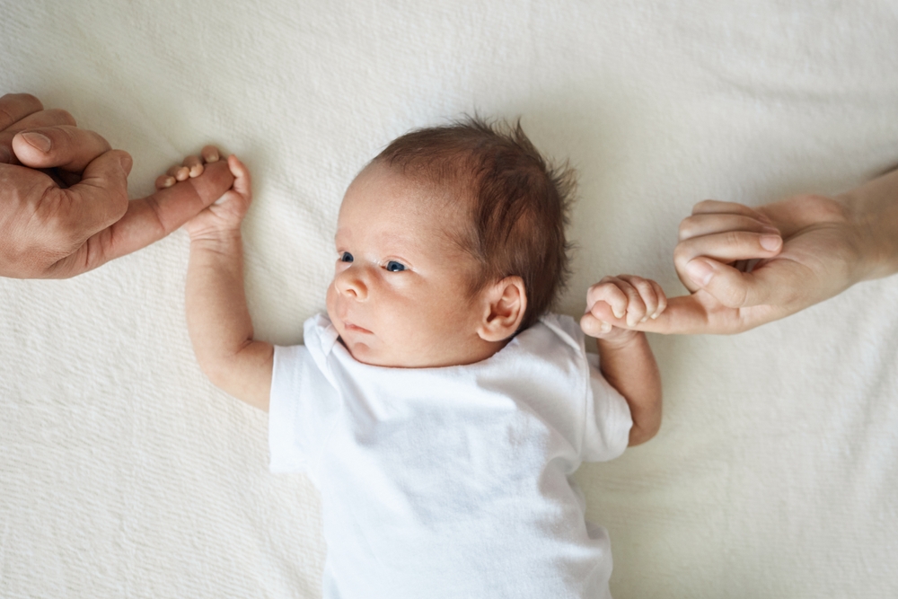 Baby holding parents hands. Father and mother hold newborn kid cute infant child in white bodysuit lying on white sheet on bed