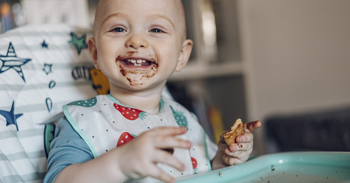 infant child sitting in high chair eating. Their face is covered in food.