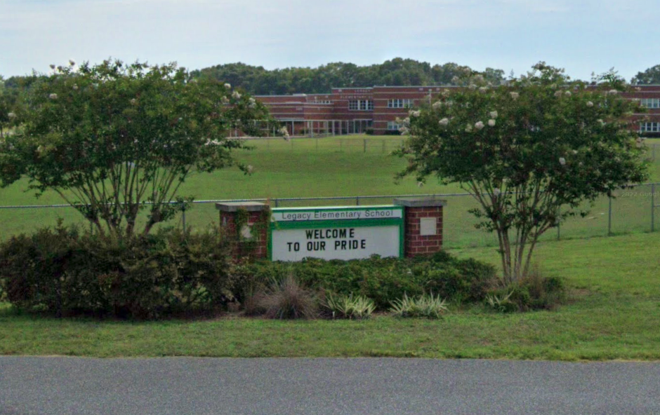 Legacy Elementary School's entrance sign reading "WELCOME TO OUR PRIDE" sits amid manicured shrubs and trees, with the brick school building visible in the distance. This is the place where over 25 students would later describe a classroom environment ruled by fear and humiliation.