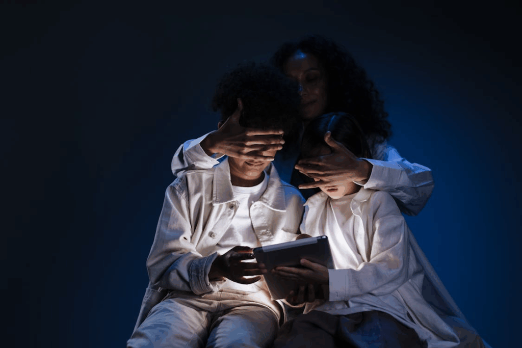 An adult with curly hair is covering the eyes of a child while they look at a tablet device in a darkened room. The image, lit with blue light from the screen, visually represents online ID check protection measures, showing parental supervision and safeguarding of children from potentially inappropriate digital content.